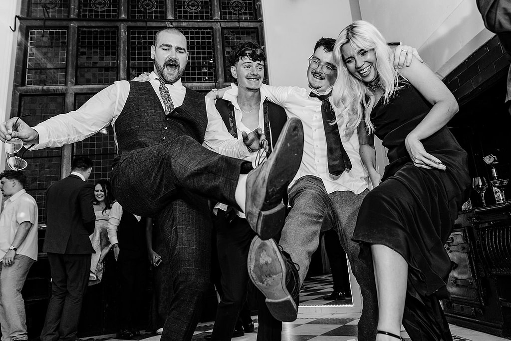 Four people dressed in formal attire laugh and kick their legs up while posing together on a chequered floor at a lively indoor event. Others mingle in the background. The scene is joyful and celebratory.
