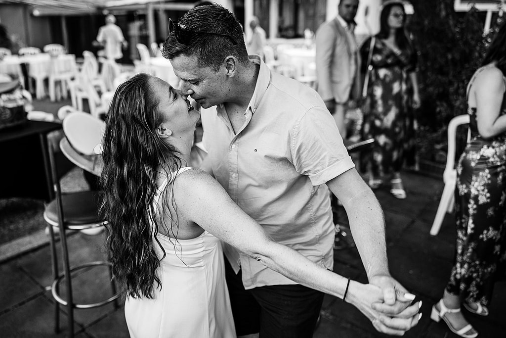 A couple dances closely outdoors, smiling and gazing into each other's eyes. Captured by a natural wedding photographer, the black and white photo features tables and guests in the background, hinting at a joyful celebration.