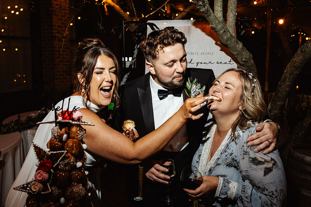At a Carcassonne wedding, a bride feeds pudding to a laughing guest while a man in a dinner jacket stands between them, smiling. Fairy lights and a decorated pudding tower shimmer in the festive, nighttime setting.