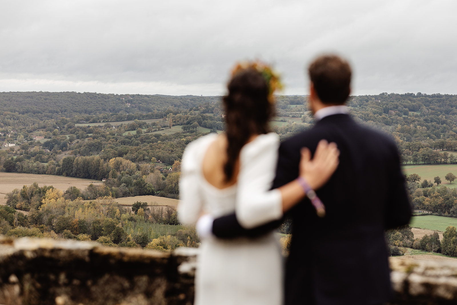 A couple, seen from behind and out of focus, stand arm in arm on a stone terrace overlooking a vast, green countryside under a cloudy sky—a perfect backdrop for a relaxed winter wedding in France.