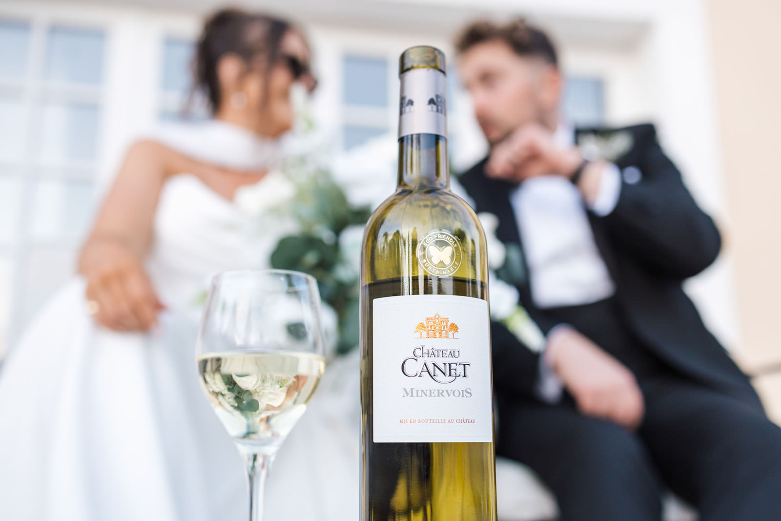 A bottle of Château Canet Minervois white wine and a filled glass are in focus in the foreground, with a bride and groom sitting together in the blurred background.