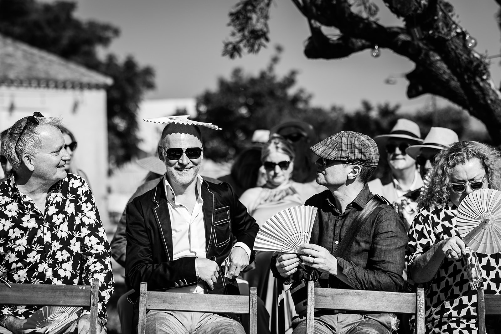 Two grooms wearing matching floral waistcoats and bow ties stand close together, holding hands and smiling at each other outdoors. The black-and-white photo radiates the joy of an intimate wedding in France, set against a soft, natural countryside background.