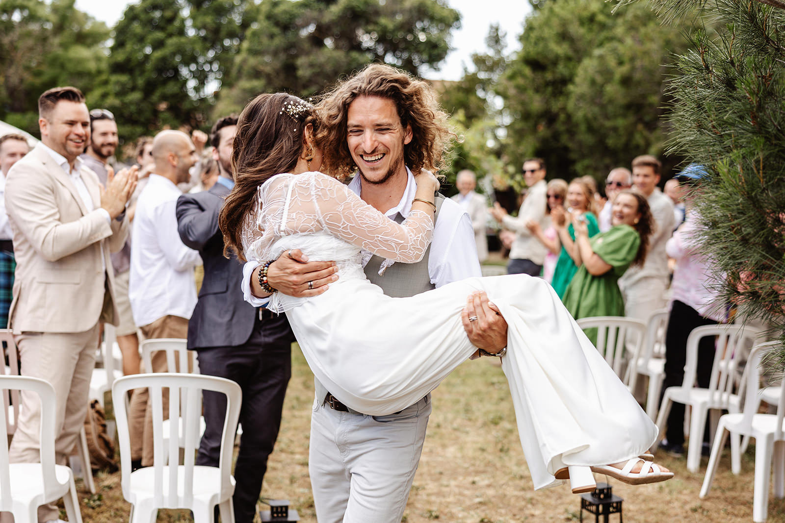 A groom with long curly hair joyfully carries his bride, who is wearing a white dress, down an outdoor aisle as guests smile and clap in celebration—captured beautifully by a south of France wedding photographer. Trees and white chairs are visible in the background.