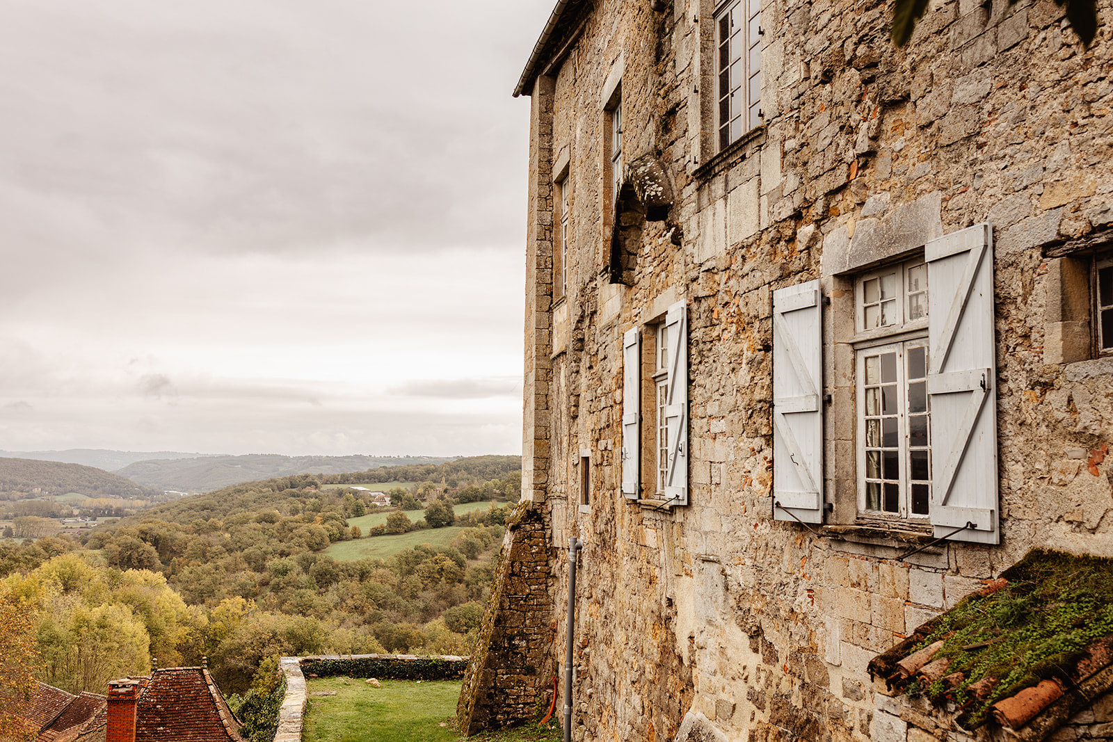A stone building with blue shutters overlooks a scenic valley of rolling green hills and distant trees under a cloudy sky, perfect for enchanting winter wedding photography in France.