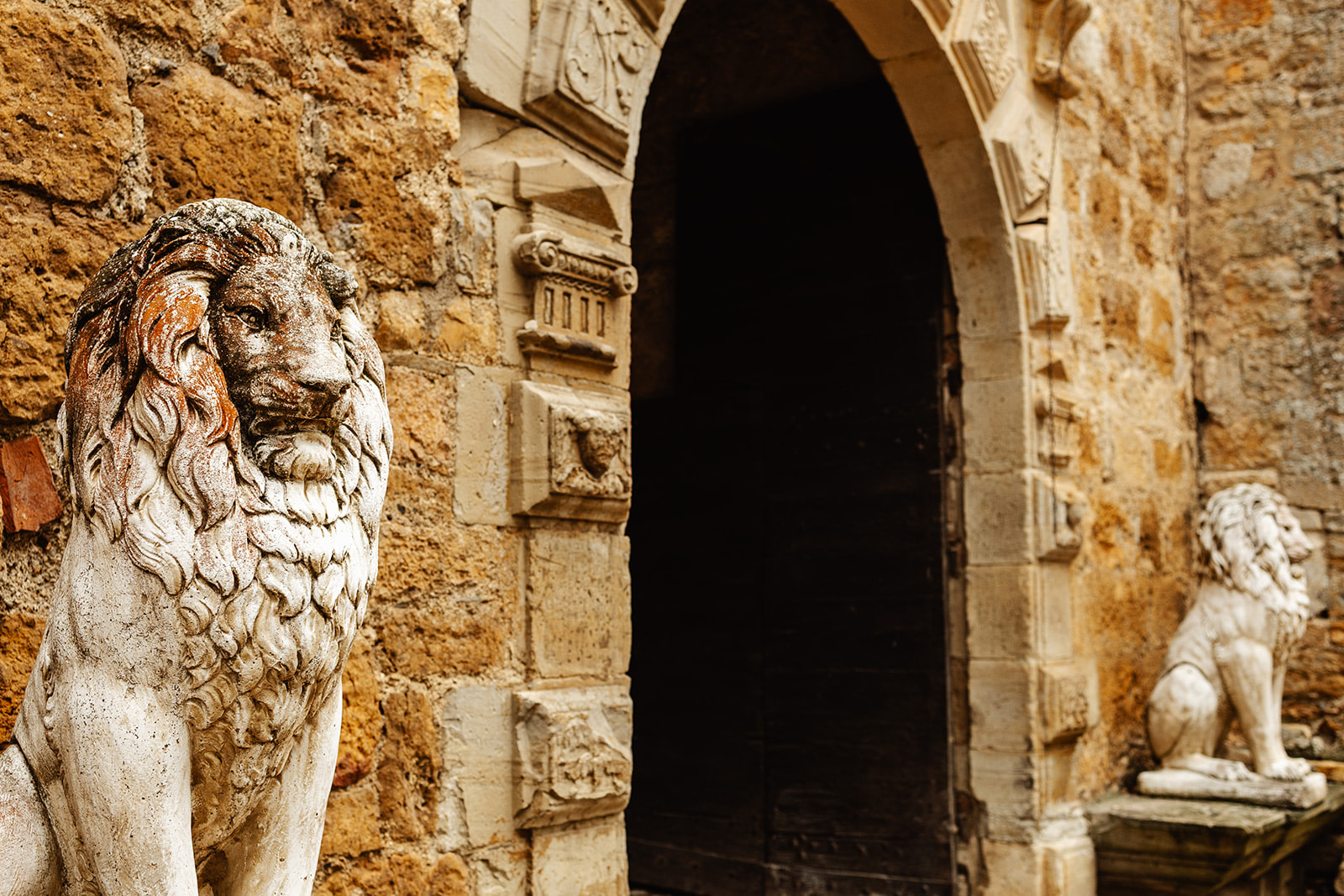 Two stone lion statues guard the entrance of an old stone building with an arched doorway in chateau-de-beduer-wedding-venue.jpg; one lion is closer and more detailed, while the other stands further in the background.
