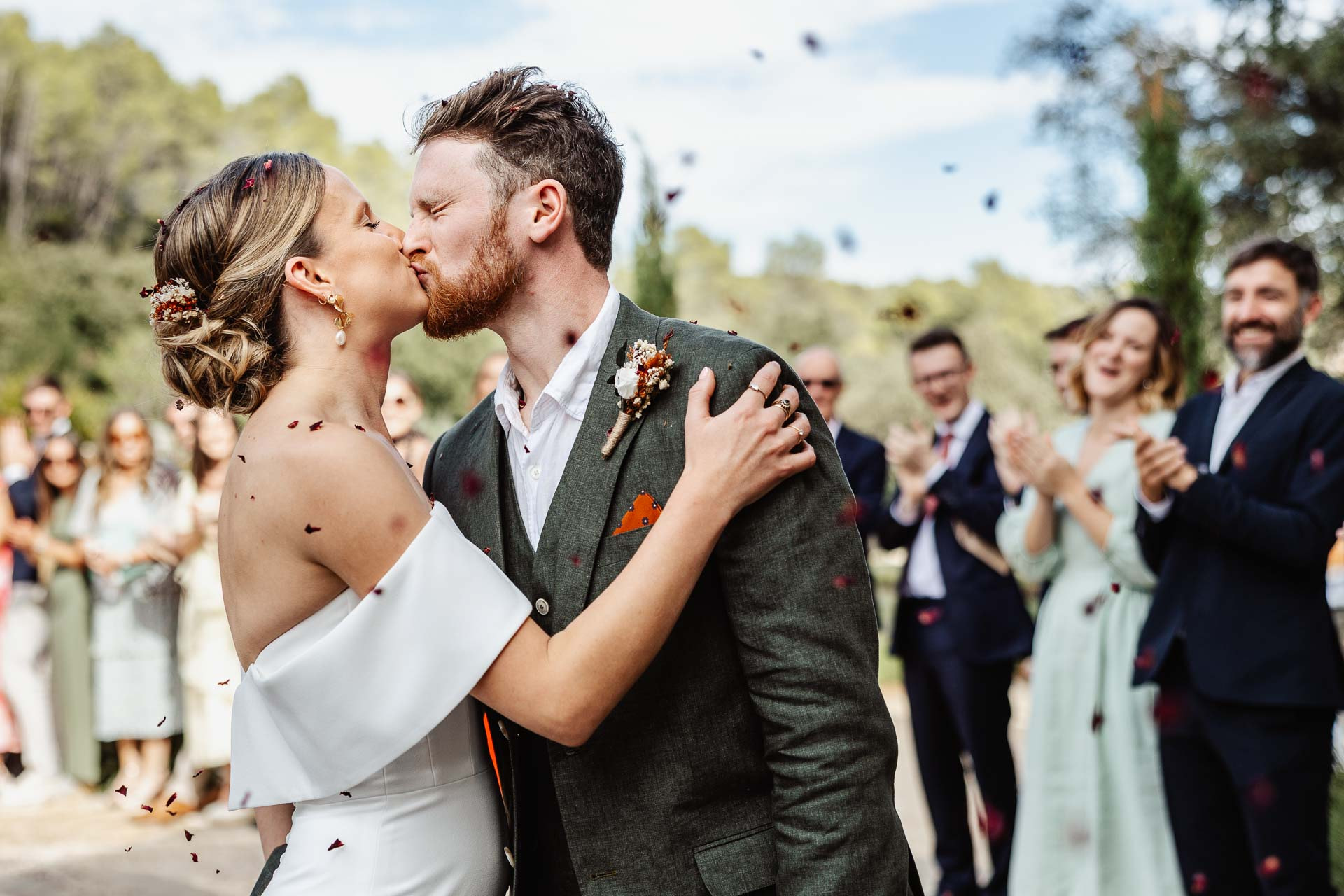 A newlywed couple kisses outdoors as confetti floats in the air, captured beautifully by a Provence wedding photographer. Cheering guests clap and smile whilst the bride in white and groom in a dark suit celebrate their special moment.