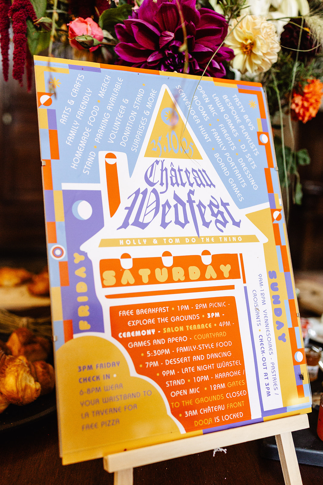 A colourful, house-shaped event schedule sign titled “Chateau Westfest” lists activities for Friday, Saturday, and Sunday. Inspired by France, the playful fonts and vibrant orange, blue, and purple evoke festive wedding photography amid flowers and food.