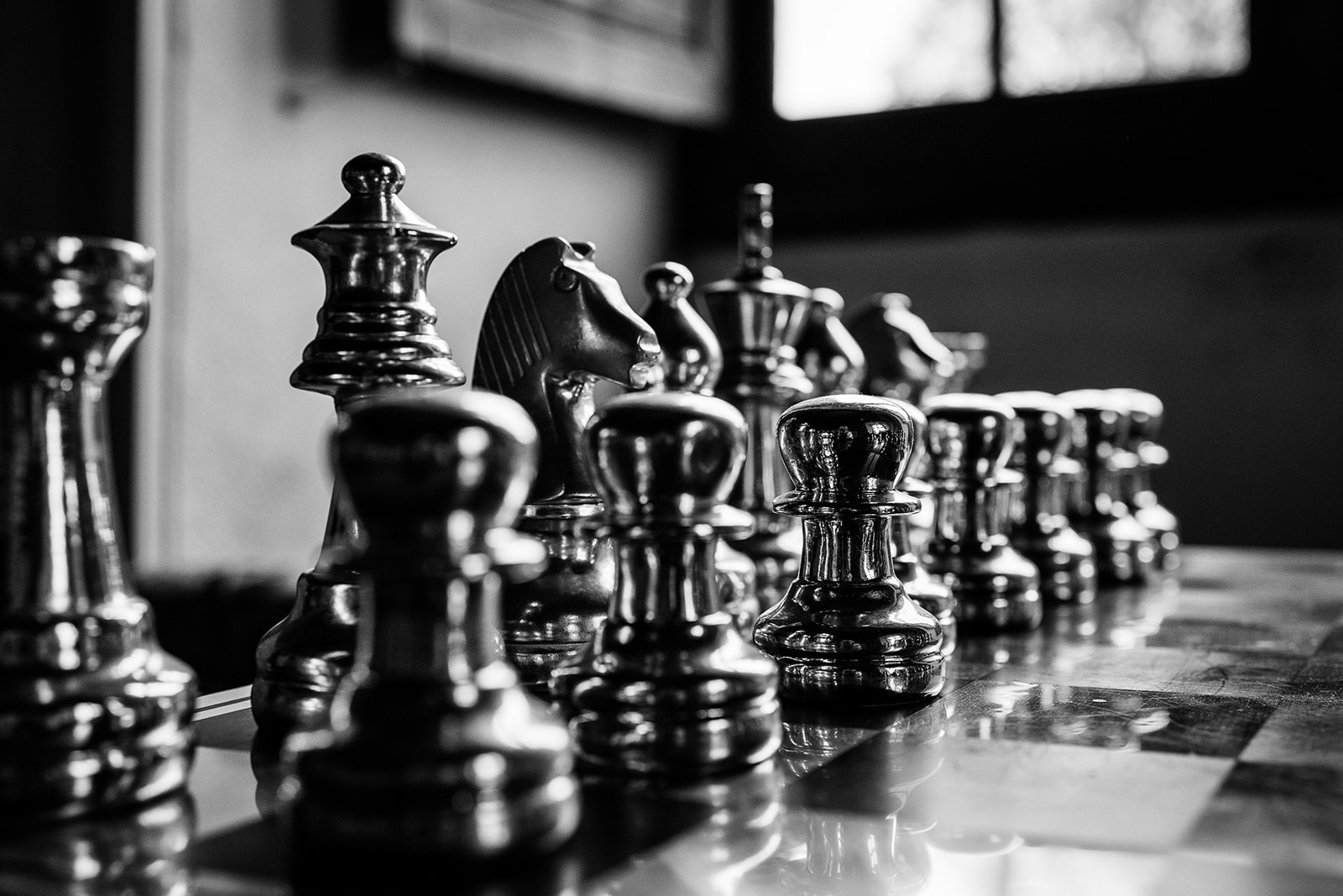A close-up, black and white photo of shiny metallic chess pieces arranged on a chessboard near a window, reminiscent of elegant wedding photography in France, with the focus on the pawns in the front row.