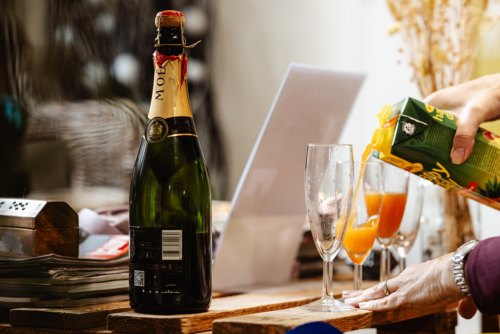 A person pours orange juice from a carton into a champagne flute beside a bottle of Moët & Chandon on a cluttered wooden table, capturing a candid moment perfect for wedding photography in France.