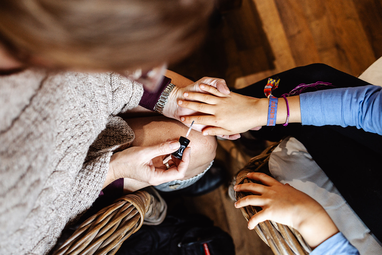 A person applies nail varnish to a child’s fingernails as the child sits in a wicker chair—another child's hand rests on the adult’s leg. The warm, casual scene with wooden floors could be a sweet moment captured during winter wedding photography in France.