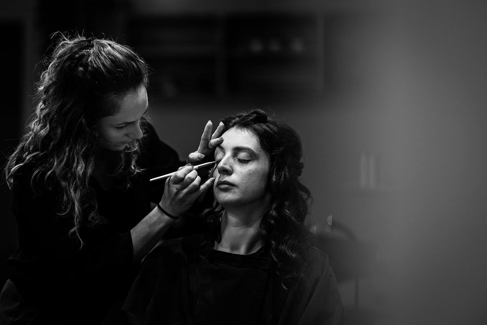 A make-up artist applies eyeshadow to a woman with her eyes closed, captured in black and white. The soft lighting and blurred background evoke the elegance of winter wedding preparations in France, perfect for wedding photography inspiration.