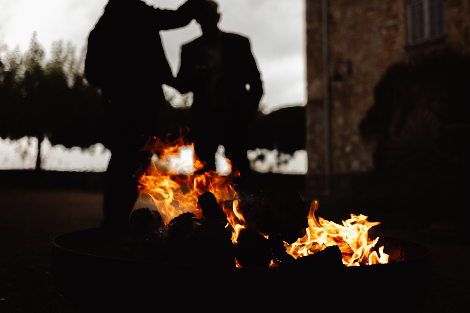 Two people are silhouetted in the background near a stone building, whilst a fire burns brightly in the foreground with visible flames and logs.