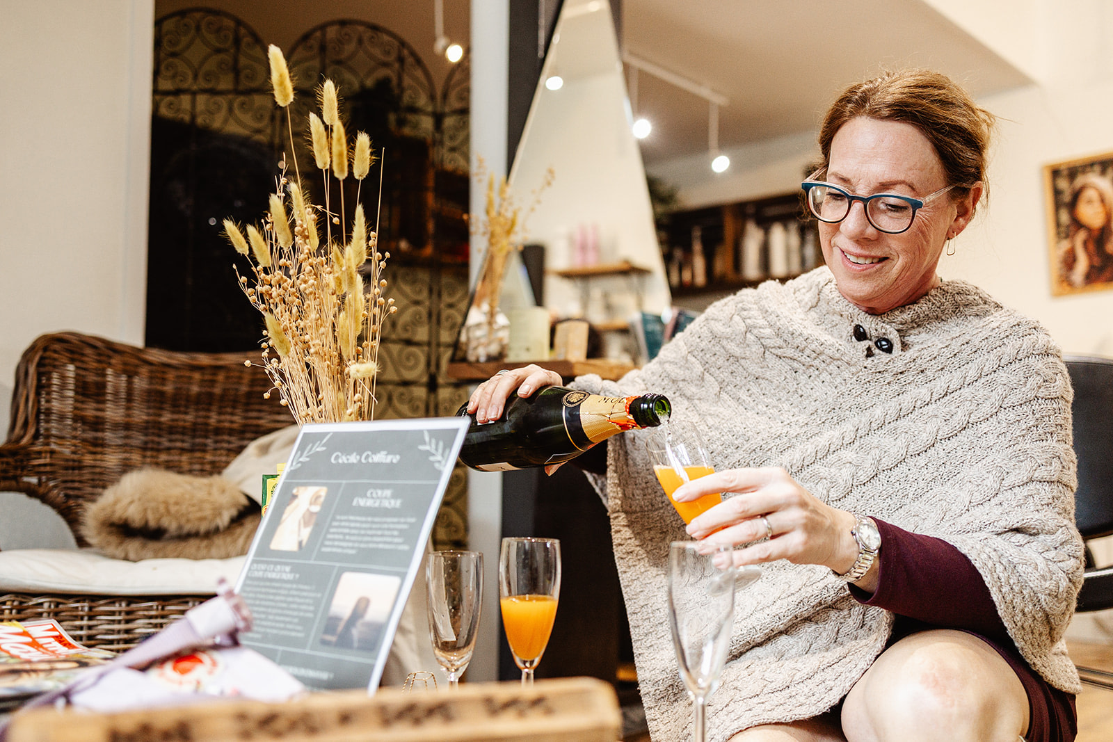 A smiling woman in glasses wearing a light knitted shawl sits indoors, pouring orange juice and champagne into a glass. On the table are magazines, dried flowers, and a café menu sign—perfect inspiration for winter wedding photography in France.