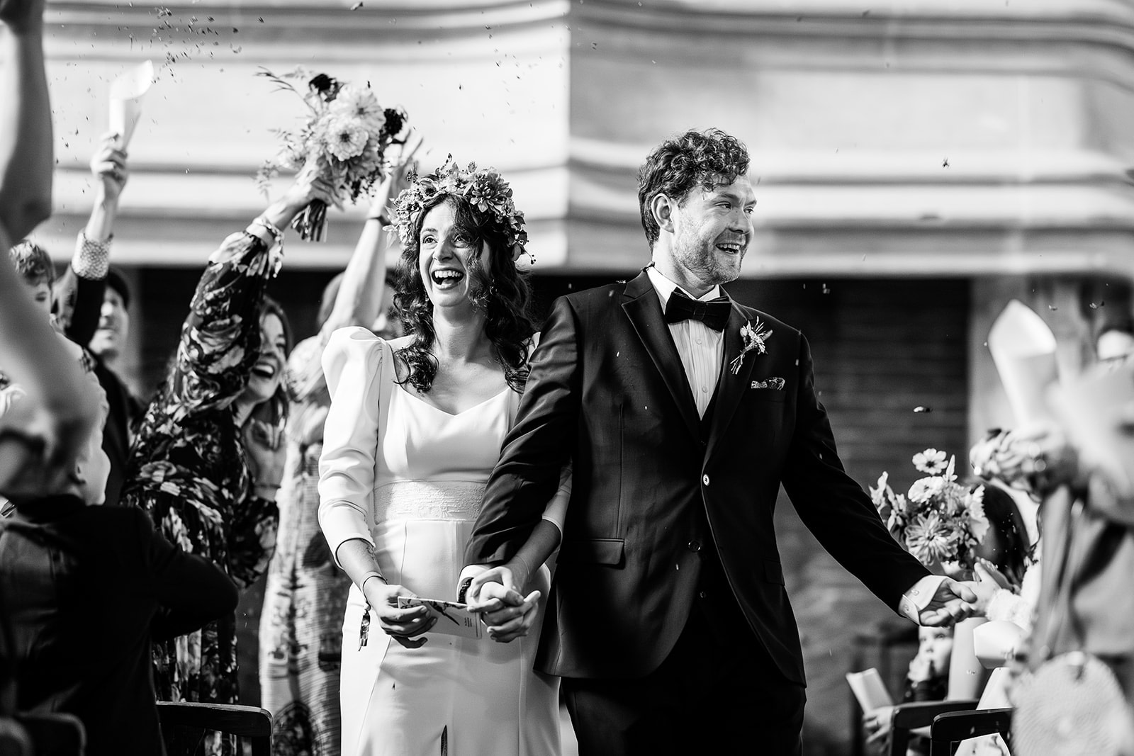 A joyful bride and groom walk hand in hand down the aisle, smiling, as guests cheer and throw confetti. The black-and-white image captures a celebratory winter wedding moment in France, perfect for wedding photography inspiration.